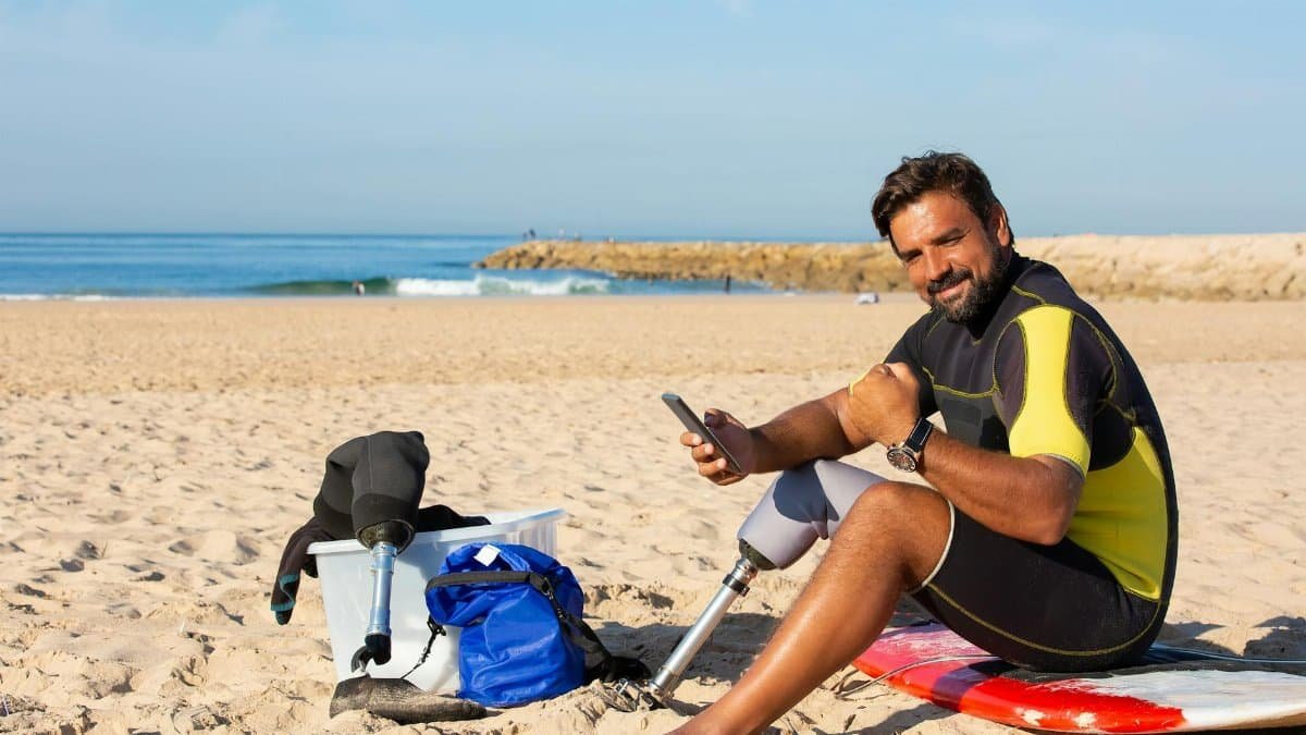 Side view full length positive ethnic male surfer with amputated leg sitting on sunny sandy beach on surfboard and browsing mobile phone while looking at camera contentedly