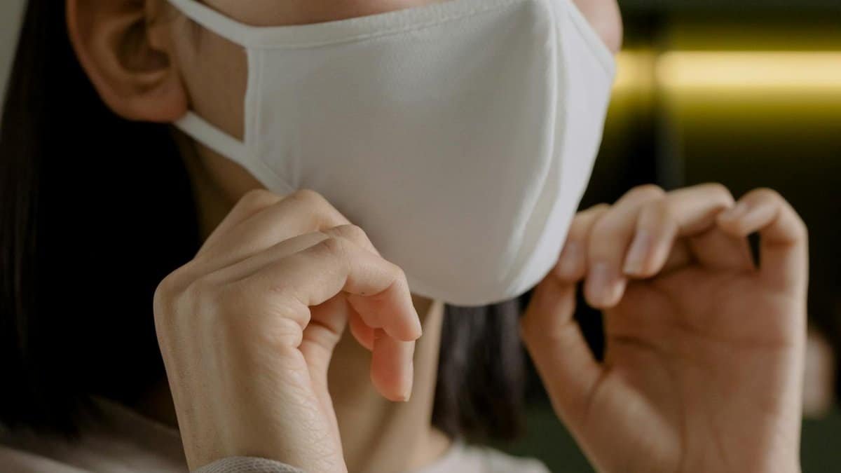 A woman adjusts her white face mask indoors, emphasizing safety and protection.