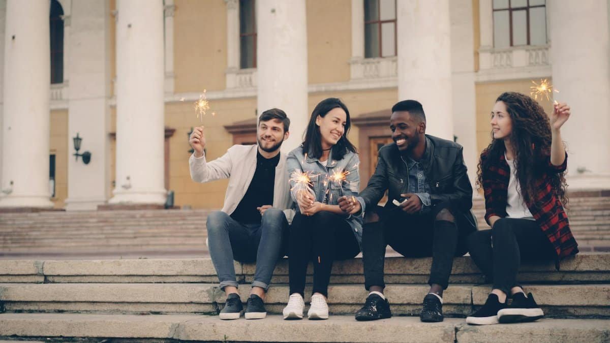 Four friends holding sparklers and sitting on steps, celebrating outdoors.