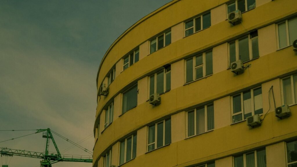 Curved yellow building facade with air conditioners and crane in urban skyline.