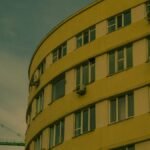 Curved yellow building facade with air conditioners and crane in urban skyline.