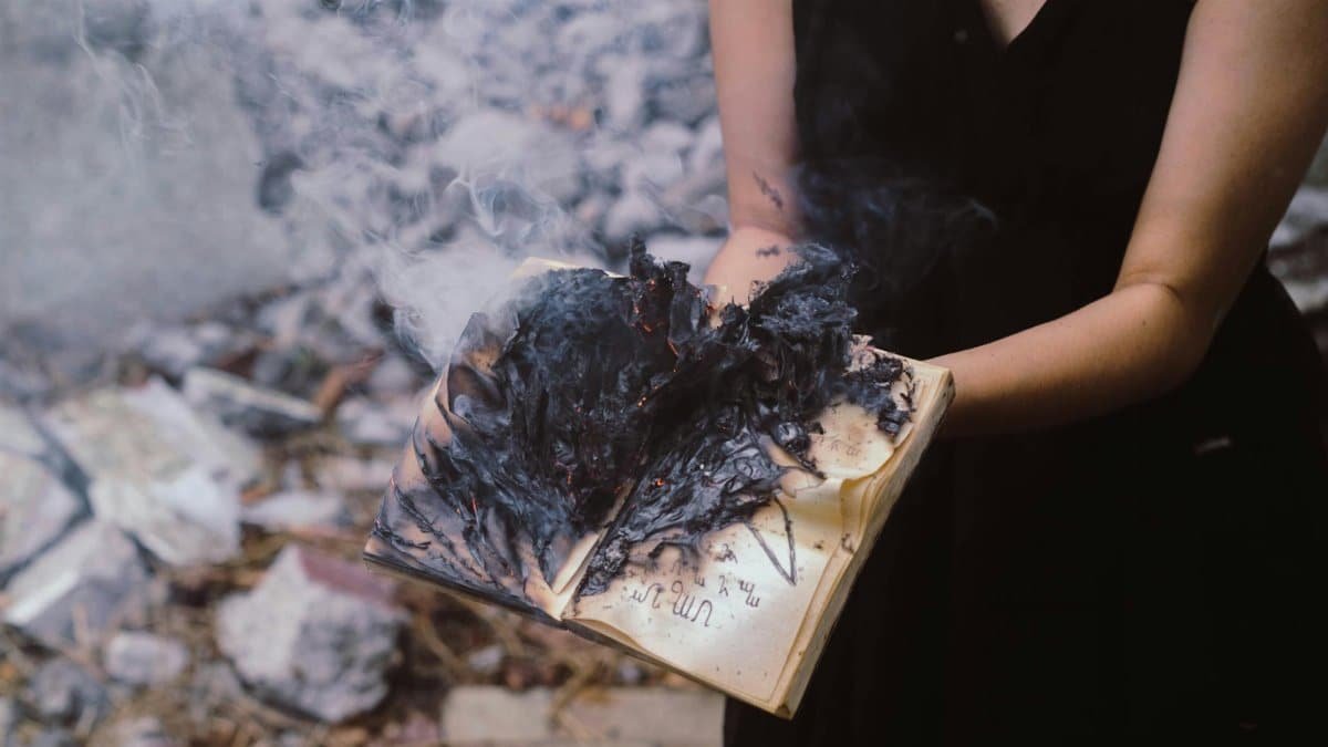 A woman holds a burning book, with smoke and ashes rising outdoors.