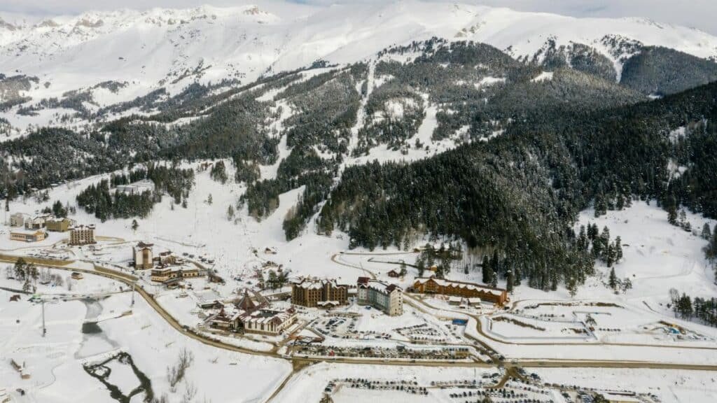 Drone shot of a snowy mountain ski resort with coniferous trees and snowy peaks.