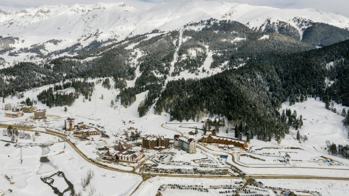 Drone shot of a snowy mountain ski resort with coniferous trees and snowy peaks.