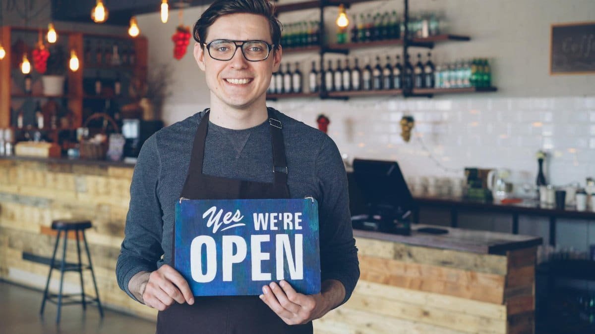 Friendly barista welcomes customers holding an open sign in a cozy cafe setting.