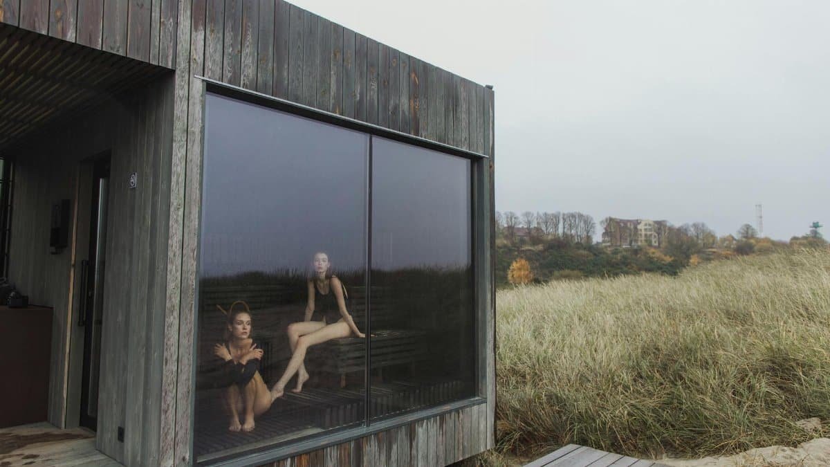 Two women in swimsuits relaxing in a wooden sauna with glass window overlooking nature.