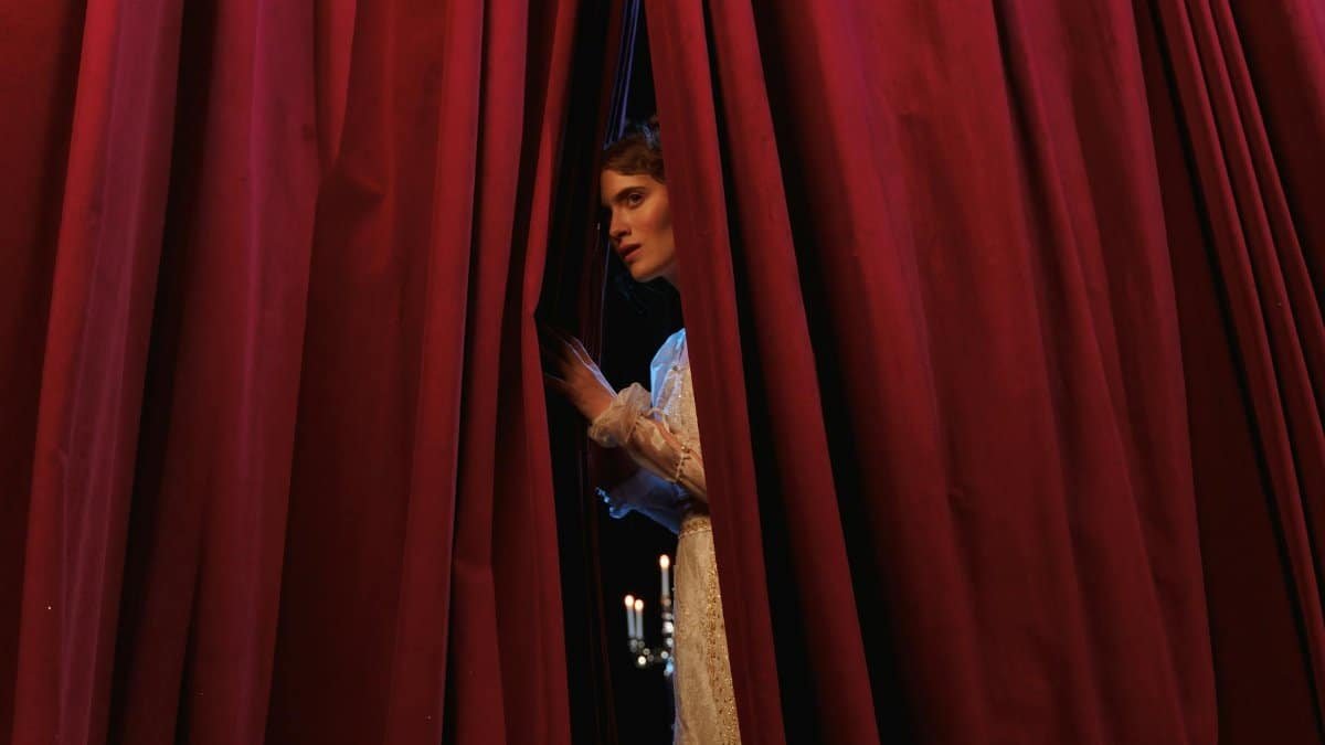 Woman peeks through red theater curtains, capturing a theatrical moment.