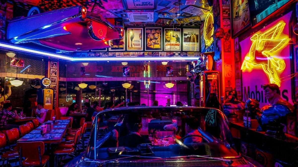 A lively retro diner bustling with people, neon lights, and classic car decor, set at night.