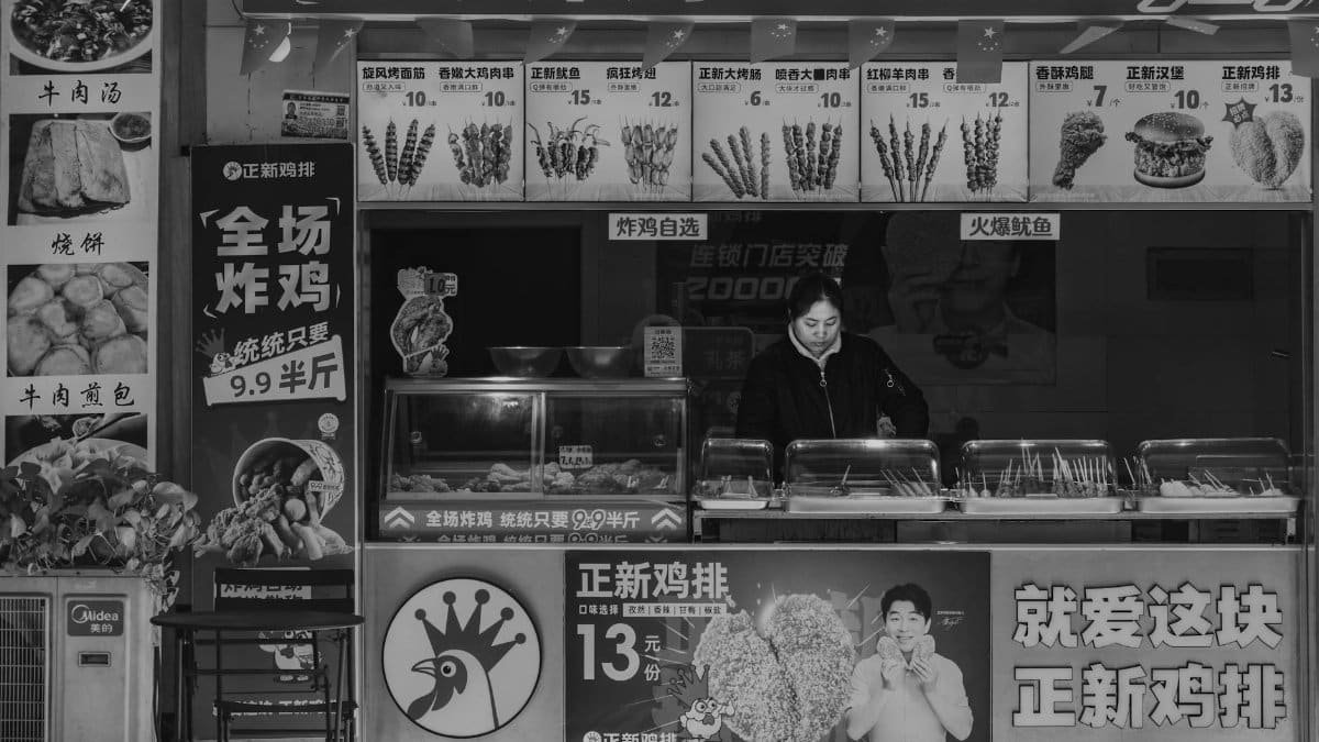 Black and white photo of a street food stall with vendor, displaying various menu items and prices.