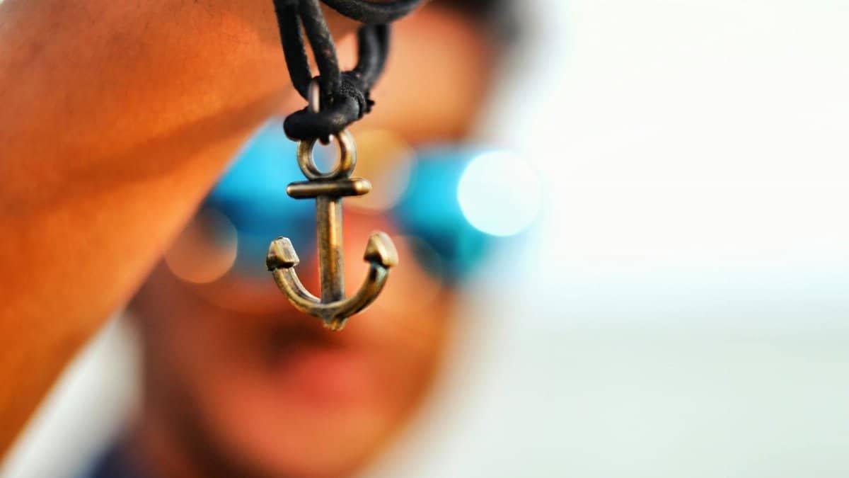 Close-up of a hand holding an anchor pendant, blurred face and sunglasses in background.
