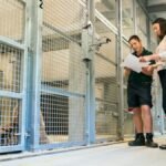 Two women reviewing plans near a tiger enclosure at a zoo, focusing on animal welfare and engineering.