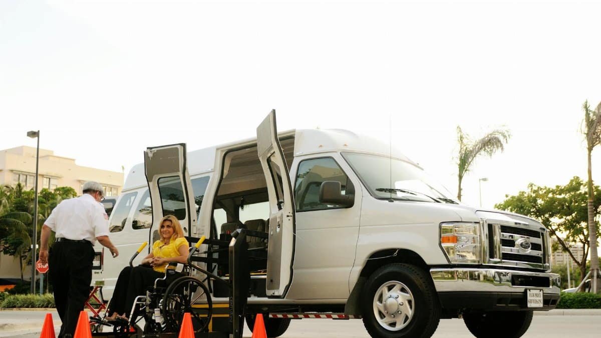 A person in a wheelchair uses a lift to board a van, assisted by a driver. Outdoor setting with cones.