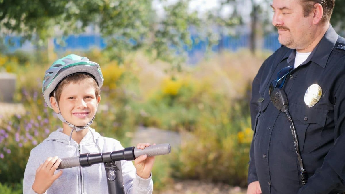 A police officer smiling at a boy on an electric scooter in a park setting, promoting community safety.