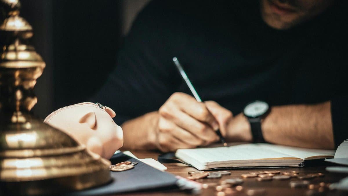 A man writing in a notebook at a desk with a piggy bank and scattered coins, suggesting financial planning.