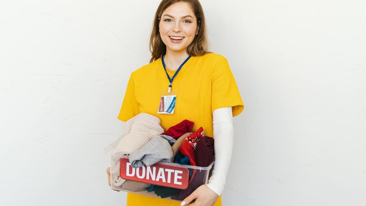Cheerful young woman in yellow shirt holding a donation box with clothes against a white background.