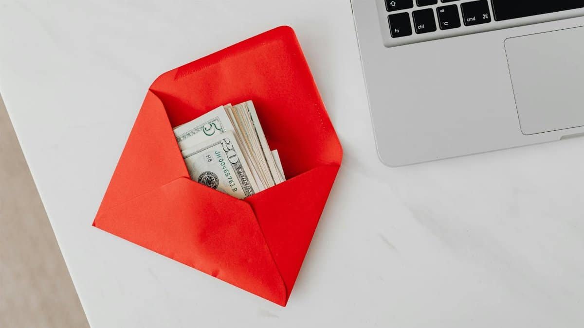 A red envelope filled with dollar bills next to a laptop on a marble desk.