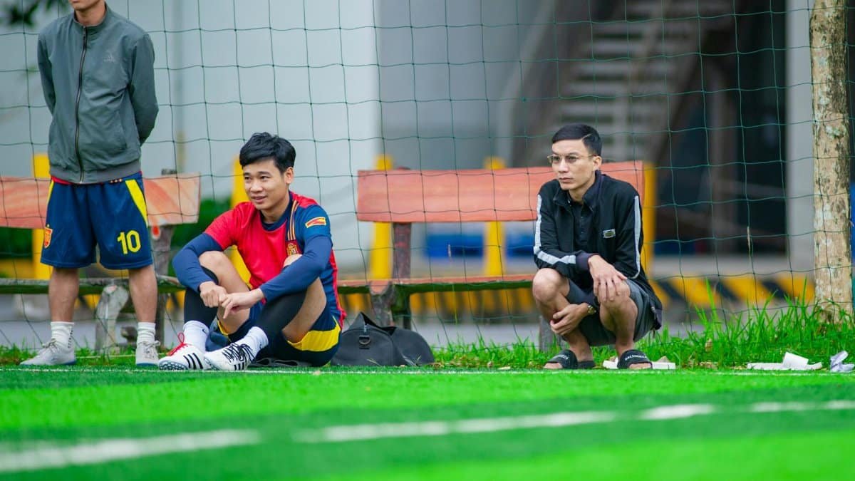 Football players sitting on the sideline of a green field in Hanoi, Vietnam.