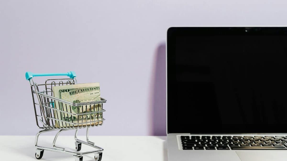 Shopping cart with money next to a laptop symbolizing online shopping and e-commerce.