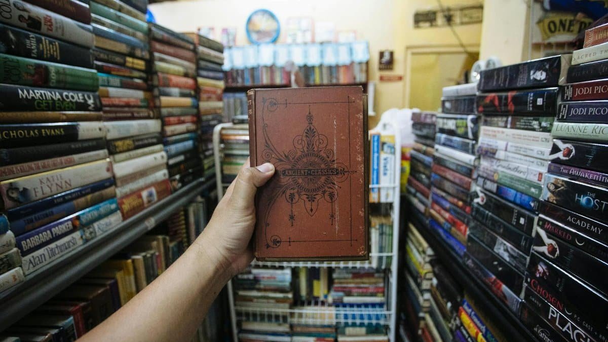 A hand holding a vintage book amidst shelves filled with books in a cozy bookstore.