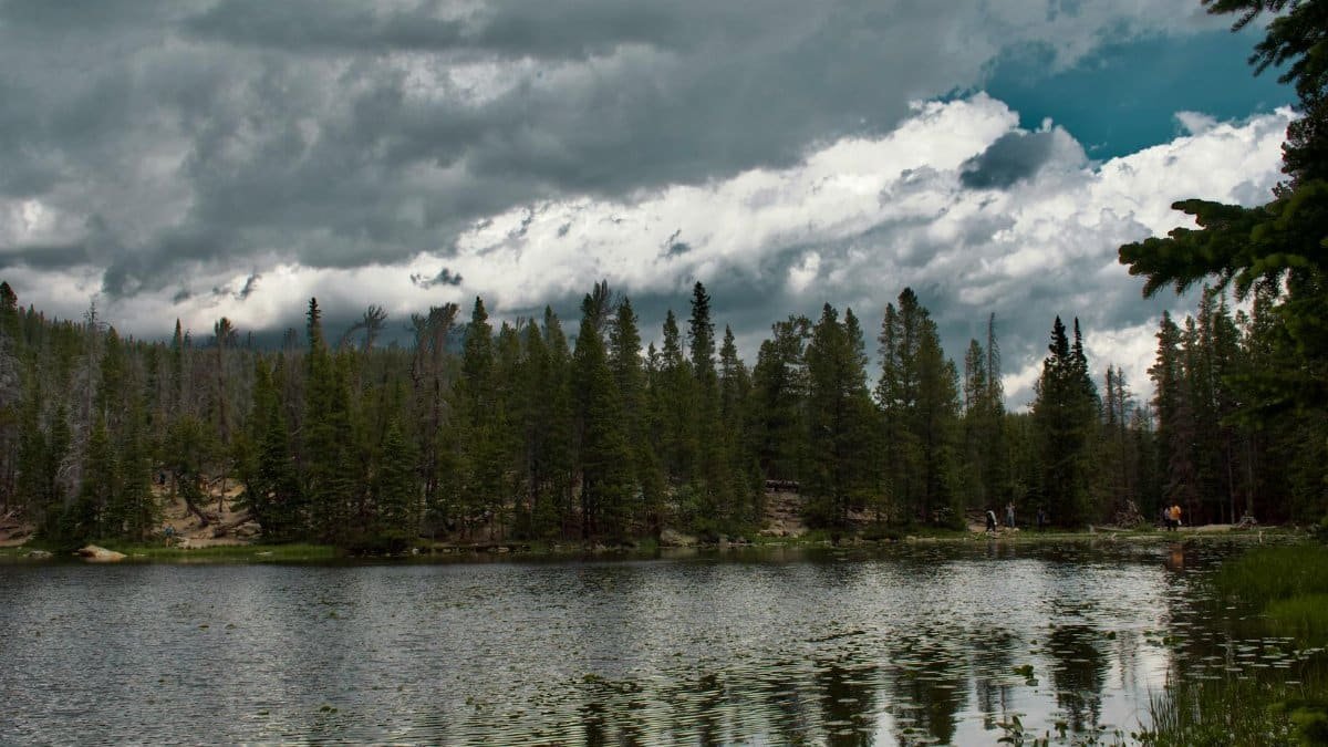 Calm lake surrounded by dense forest under dramatic storm clouds, creating a peaceful yet moody atmosphere.