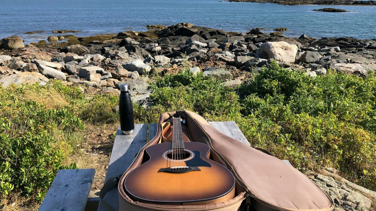 Acoustic guitar in case placed on wooden table on stony coast washed by blue rippling sea on sunny summer day
