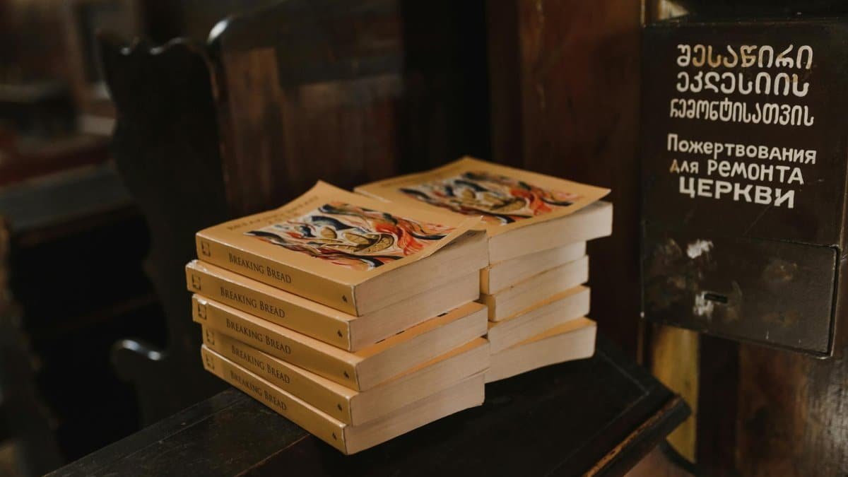 A stack of books titled 'Breaking Bread' on a wooden church surface with donation box.