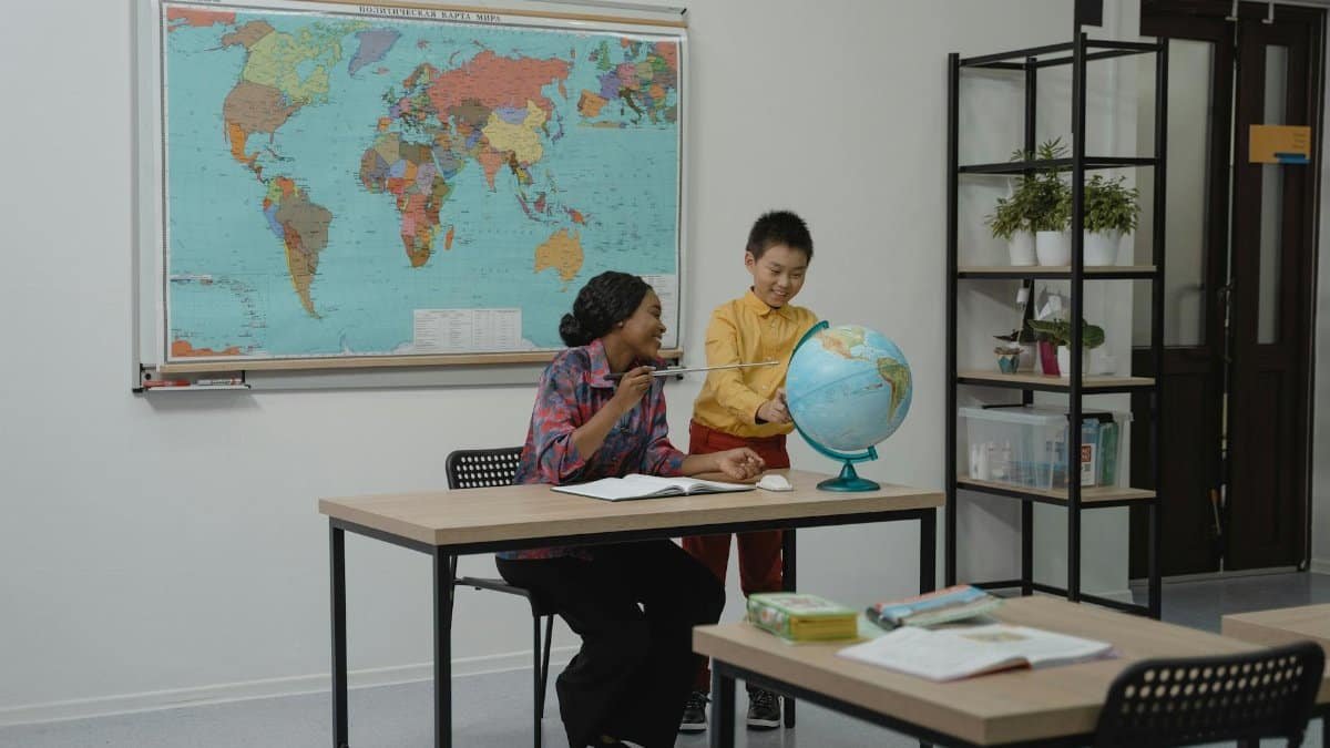 A teacher and student looking at a globe in a modern classroom setting, with a world map on the wall.