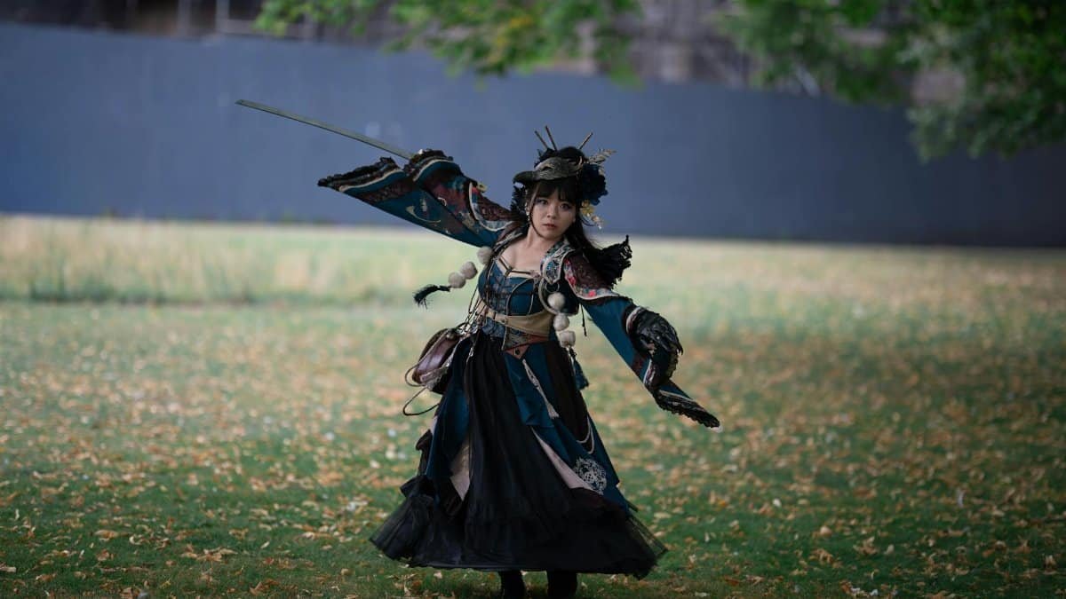 A young woman in a traditional warrior costume stands outdoors, holding a sword with a poised stance.