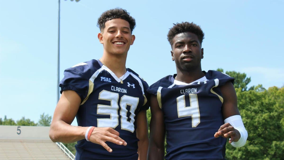 Two young football athletes posing in team uniforms outdoors on a sunny day.