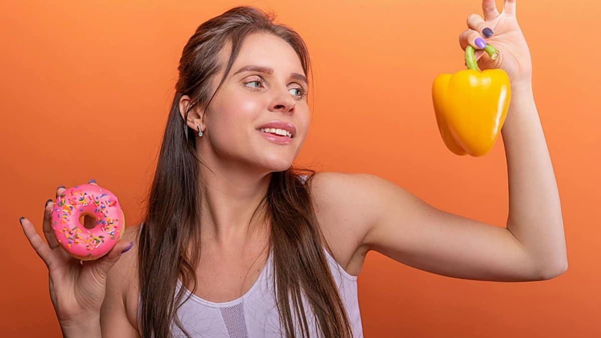 Woman in sports bra juggling choice between a donut and bell pepper on orange background.