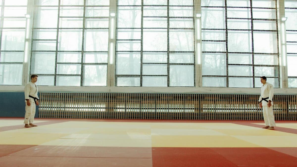Two martial artists face off in a sunlit dojo, wearing traditional judo gear.