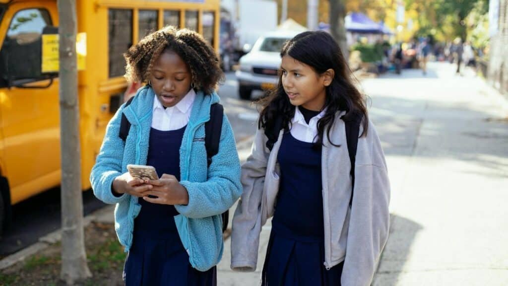 Cheerful multiracial girls classmates with backpacks browsing smartphone while walking together on sunny autumn street near yellow school bus