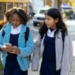 Cheerful multiracial girls classmates with backpacks browsing smartphone while walking together on sunny autumn street near yellow school bus