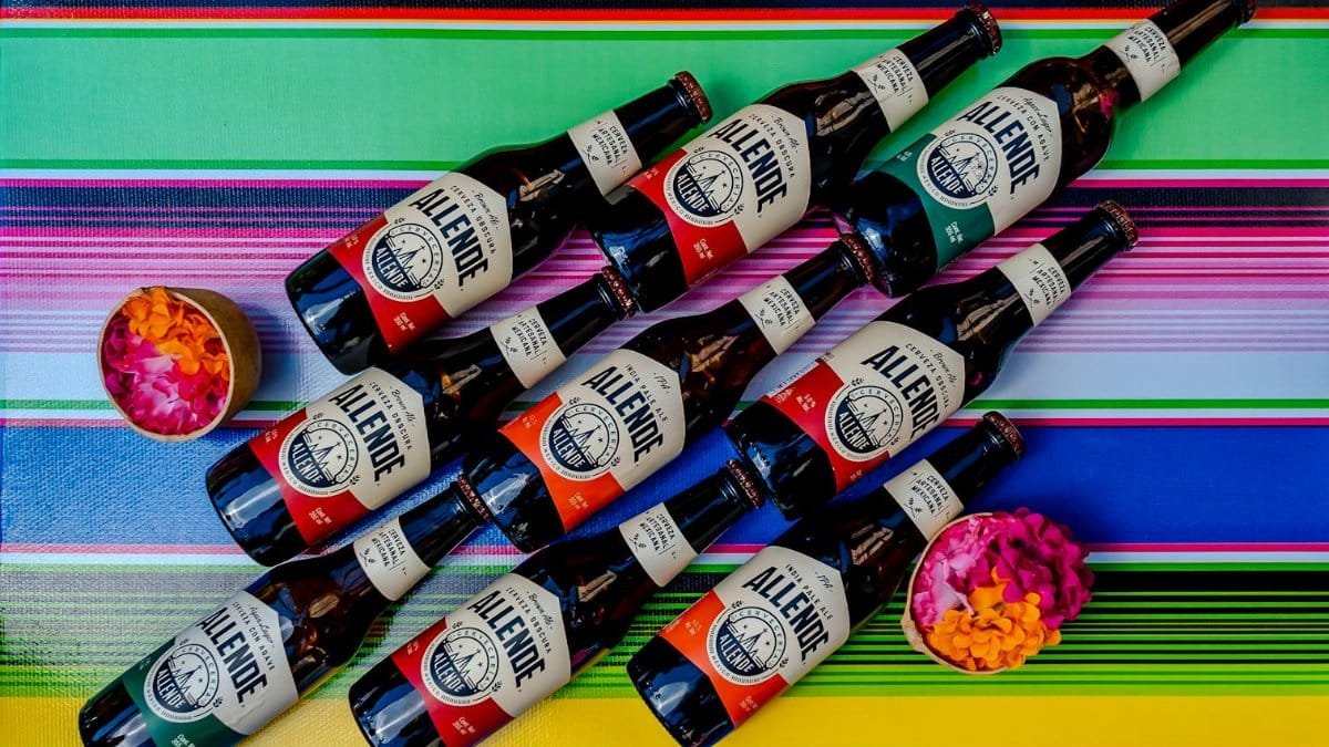 A vibrant display of Allende beer bottles on a colorful Mexican textile with flower petals.