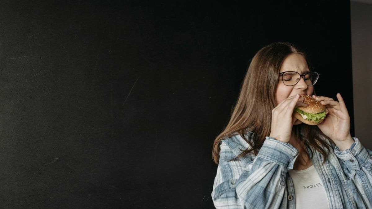 Woman in casual attire eating a hamburger in front of a dark background.