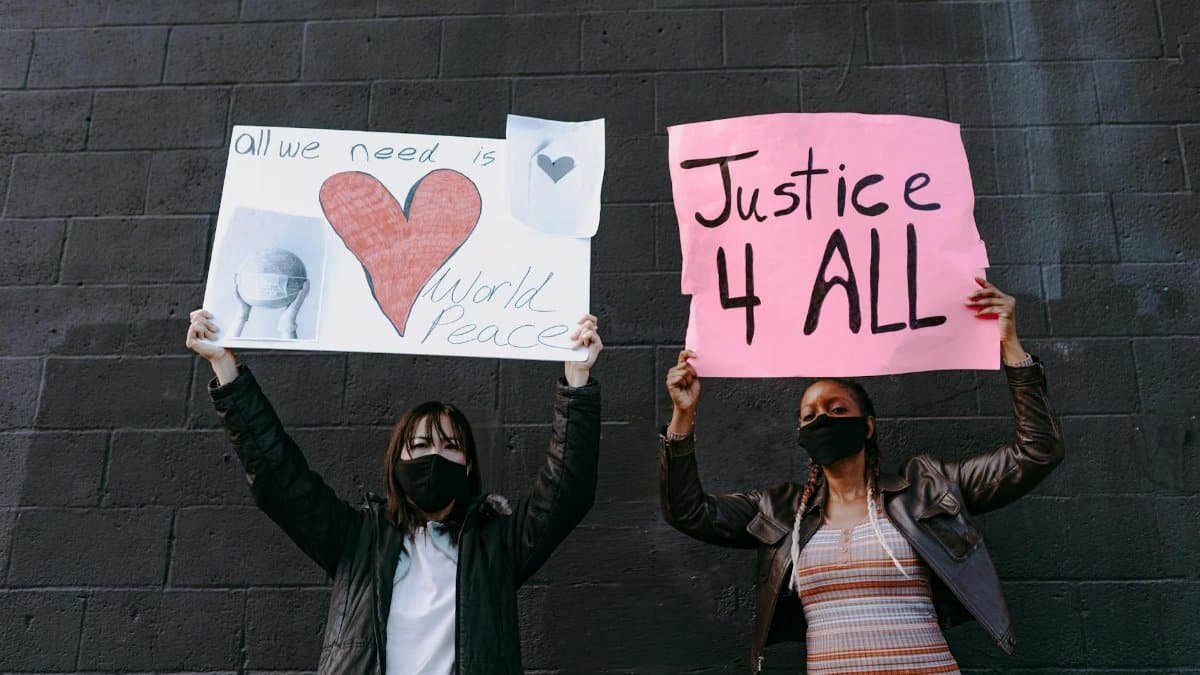 Two masked women holding protest signs advocating world peace and justice against a dark wall.