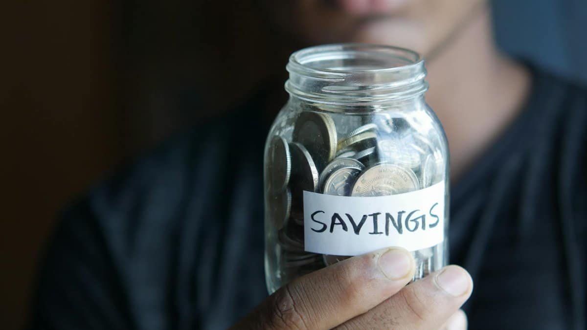 A close-up image of a person's hand holding a jar full of coins labeled 'Savings'.