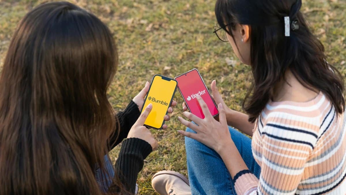Two women using dating apps Bumble and Tinder on smartphones in a park setting.