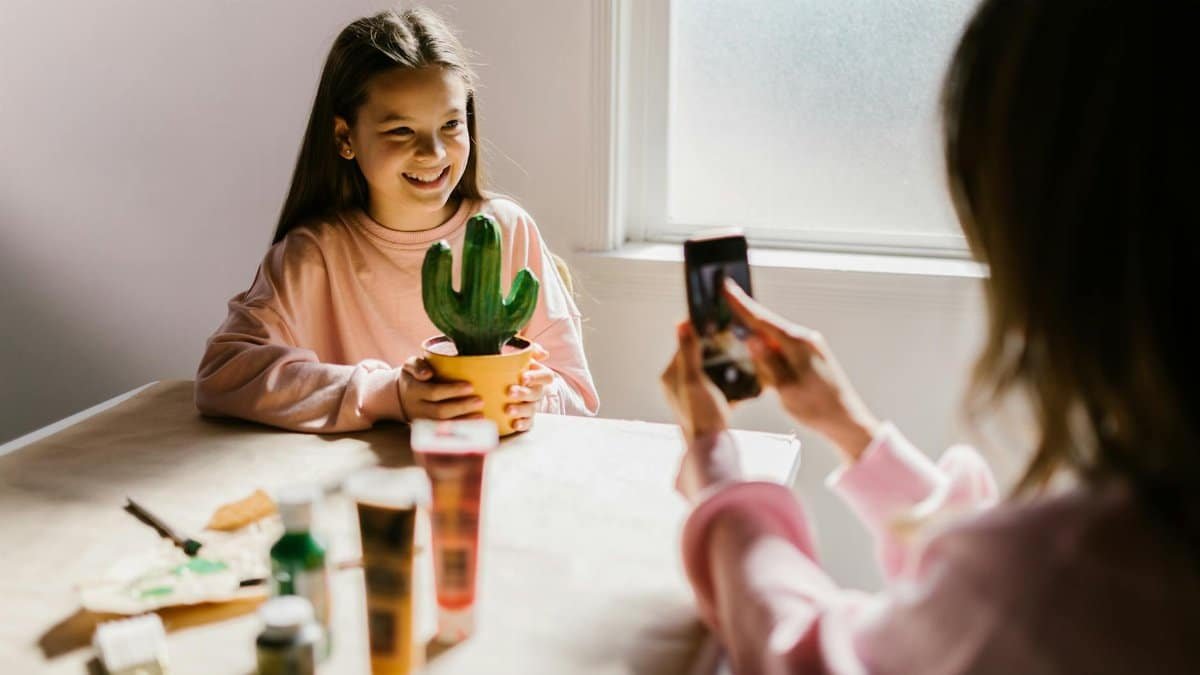 A mother photographs her daughter holding a cactus during a crafting session at home.