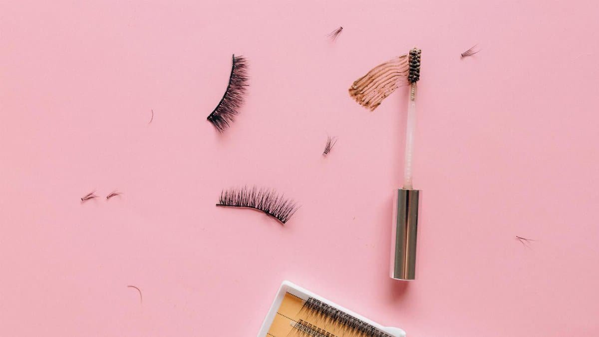 Styled flat lay of false eyelashes and makeup brush on a pastel pink surface.