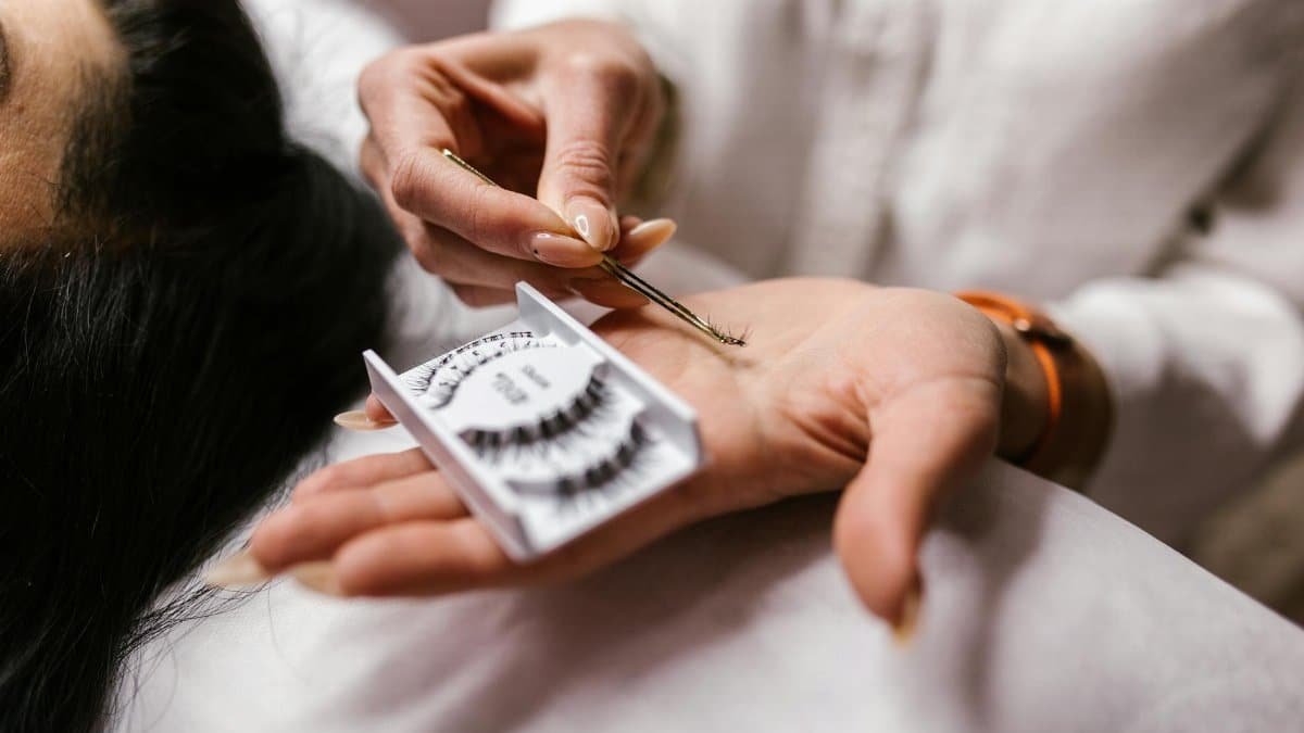 Close-up of an eyelash extension application using tweezers, featuring a beautician's hands.