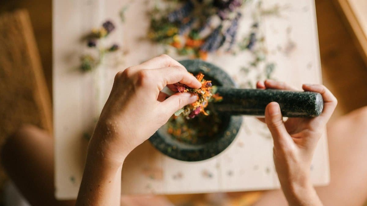 A close-up view of hands grinding herbs and flowers in a mortar and pestle on a wooden table.
