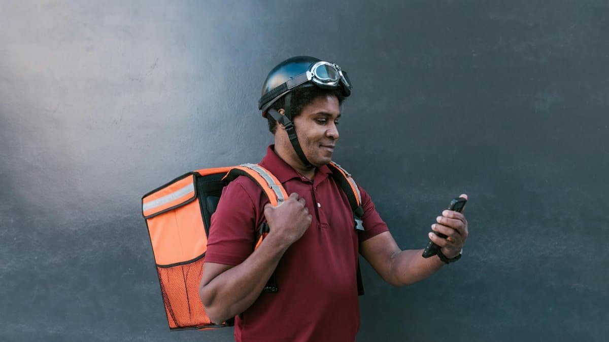 A delivery courier wearing a helmet and backpack checks his smartphone against a dark background.