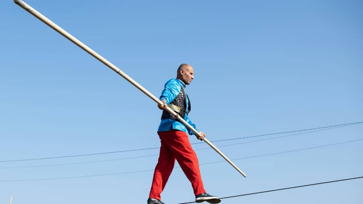 Circus performer walking on a tightrope with a balancing pole against blue sky.