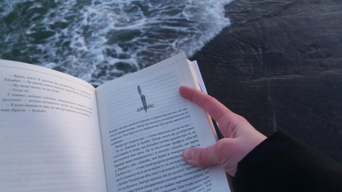 A person holds and reads a book by the ocean, enjoying a peaceful moment by the water.