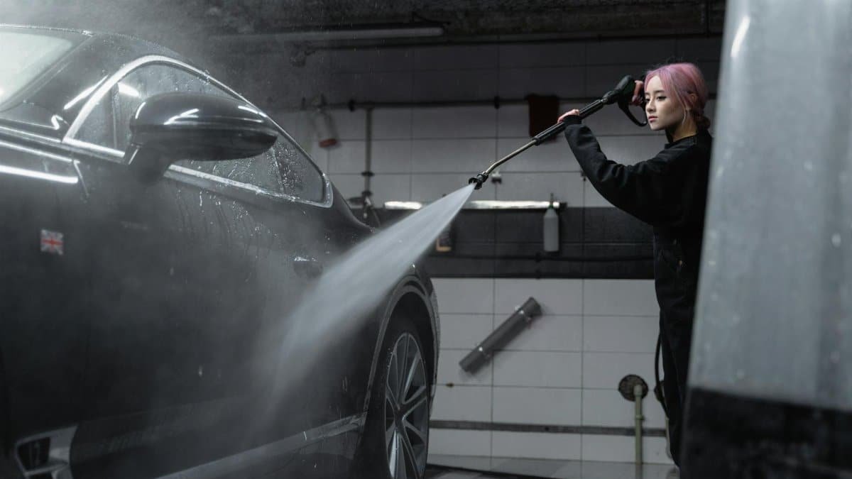 Woman power washing a black car in an indoor garage using a high-pressure washer.