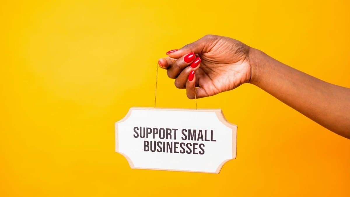 Hand holding a 'Support Small Businesses' sign against a vibrant yellow background, promoting local commerce.