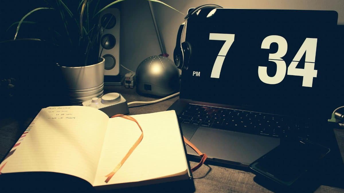 A cozy workspace with a laptop displaying time, open notebook, and headphones under warm lighting.