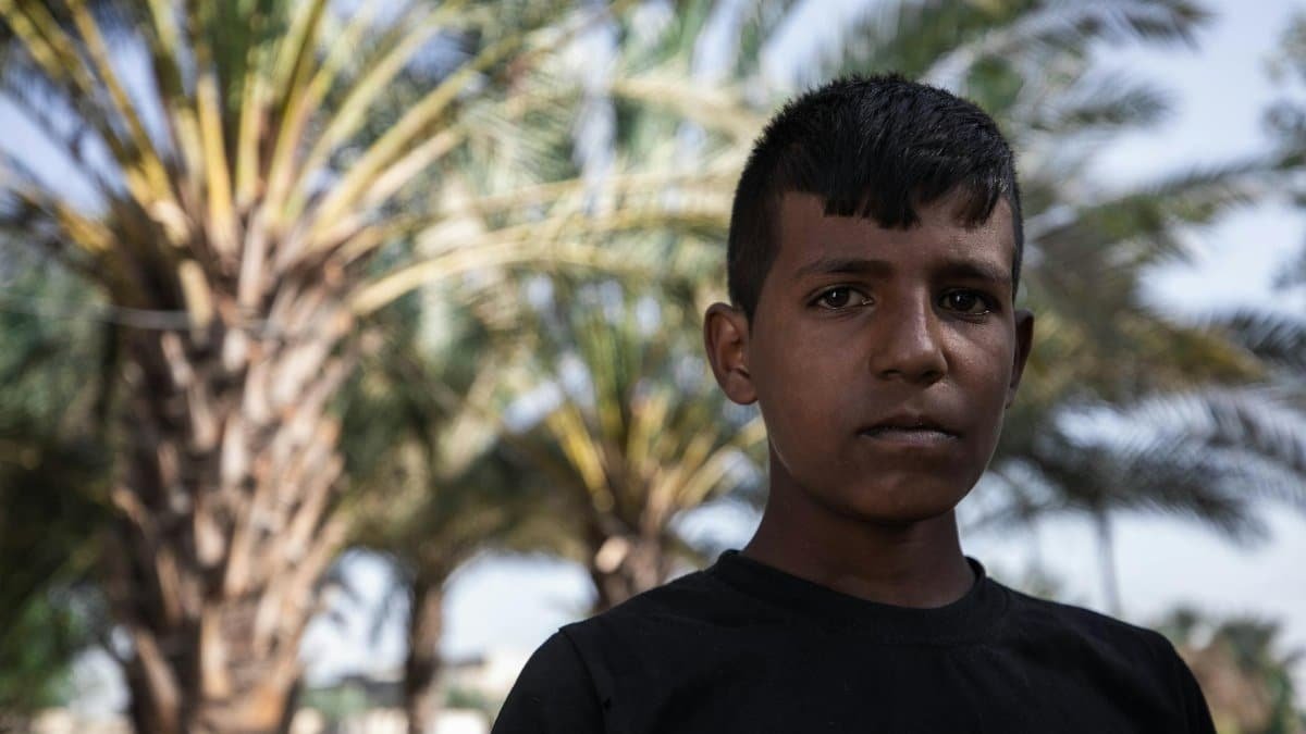 Young boy standing outdoors with palm trees in the background, looking thoughtful.
