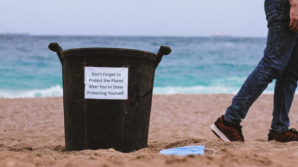 Beach scene with a trash can urging environmental protection and a discarded face mask.